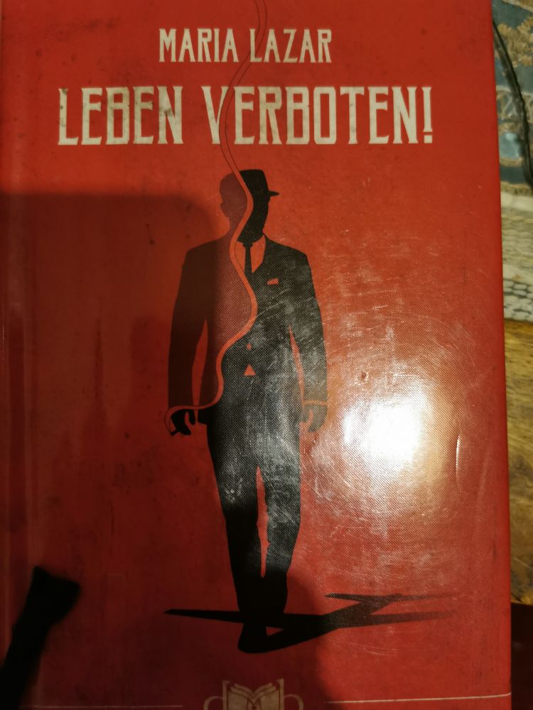 Ein Buch mit der der schwarzen, gezeichneten Figur eines Mannes mit Hut und Anzug. Der Hintergrund ist in rot gehalten. Oben der Name der Autorin Maria Lazar, darunter der Titel Leben verboten. 