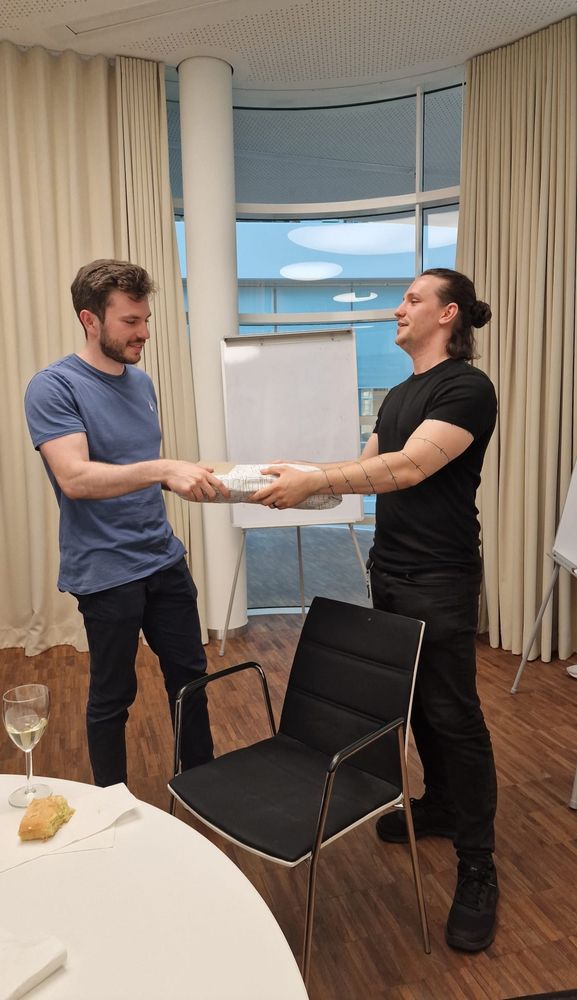 Tim receiving a farewell gift from Eugen. Both are standing in a meeting room, smiling and holding the gift between them.