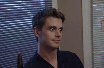 An individual stares forward eyes wide in shock. There is blinds behind them. Their hair is dark brown and short.