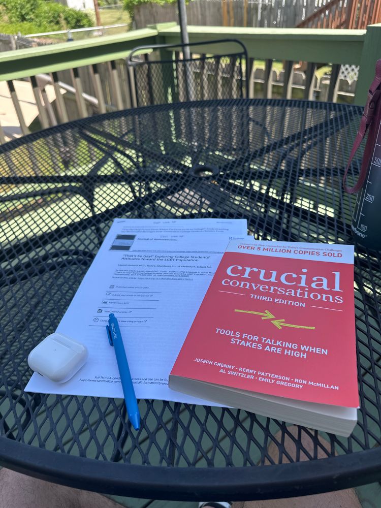 Picture of a book called Crucial Conversations, two research articles, a pen and AirPods sitting on a table.