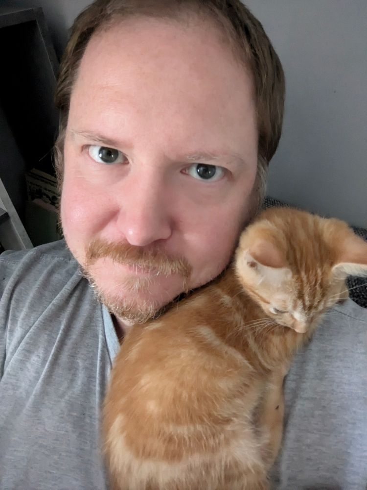 Selfie photo of a middle aged man, with a small ginger kitten curled up on his shoulder.