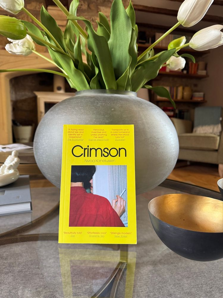 Book cover. ‘Crimson’ by Nivaq Korneliussen. This UK paperback translation has a vivid acid yellow cover, central is a square photograph. The photograph shows the partial back view of a woman. Her black hair, the white skin of her neck and her shoulder in a red shirt or dress, in her raised hand, she holds a lit cigarette. The woman appears to be standing by a door frame, from which the nose, mouth and chin of another person can be seen. 

The top of the cover has the quotes: “A fizzing read about sex and identity set in Greenland” The Economist, “Ferocious, inventive and unlike anything I’ve read” Sophie Mackintosh and “Transports us to a cold homeland where the blood runs hot” Guardian. 

Below the this is the books title “Crimson” and the author’s name. Below the photograph are more quotes: “Beautifully told” Elle, “Effortlessly Cool” Sharlene Tea, “Strikingly modern” New Yorker