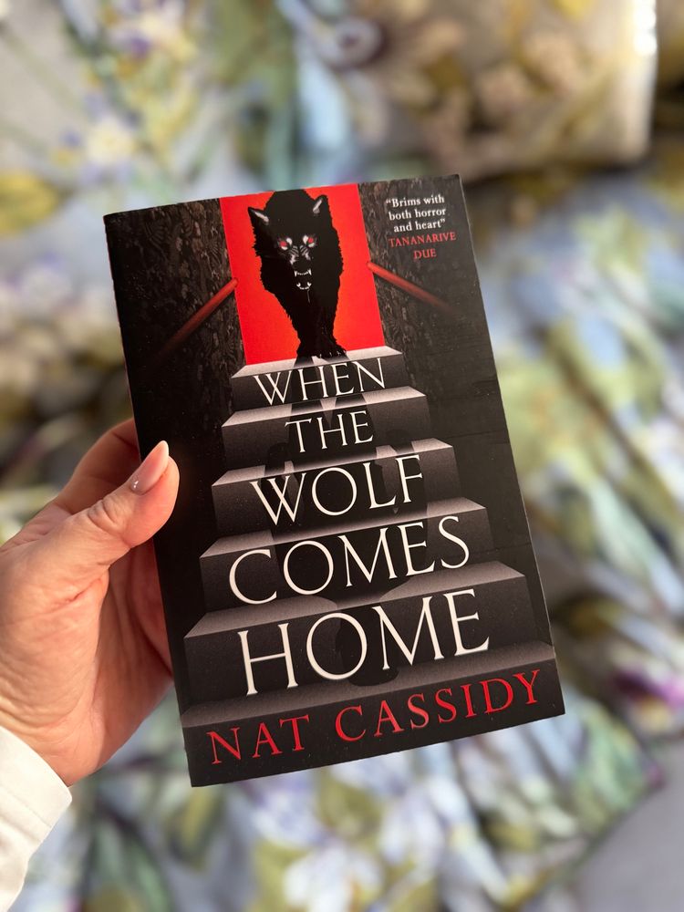 Photograph of me holding a book. This is the UK paperback edition of Nat Cassidy’s ‘When the Wolf Comes Home’. The cover is predominantly black; a drawing of a wolf, with glowing red eyes and an open fanged mouth stands within a red box/doorway, below the wolf in grey tones are descending steps, the title in white font is written one word to each step, ending with the authors name in red text. 
At the top right is a quote from Tananarive Due “Brims with both horror and heart”. 