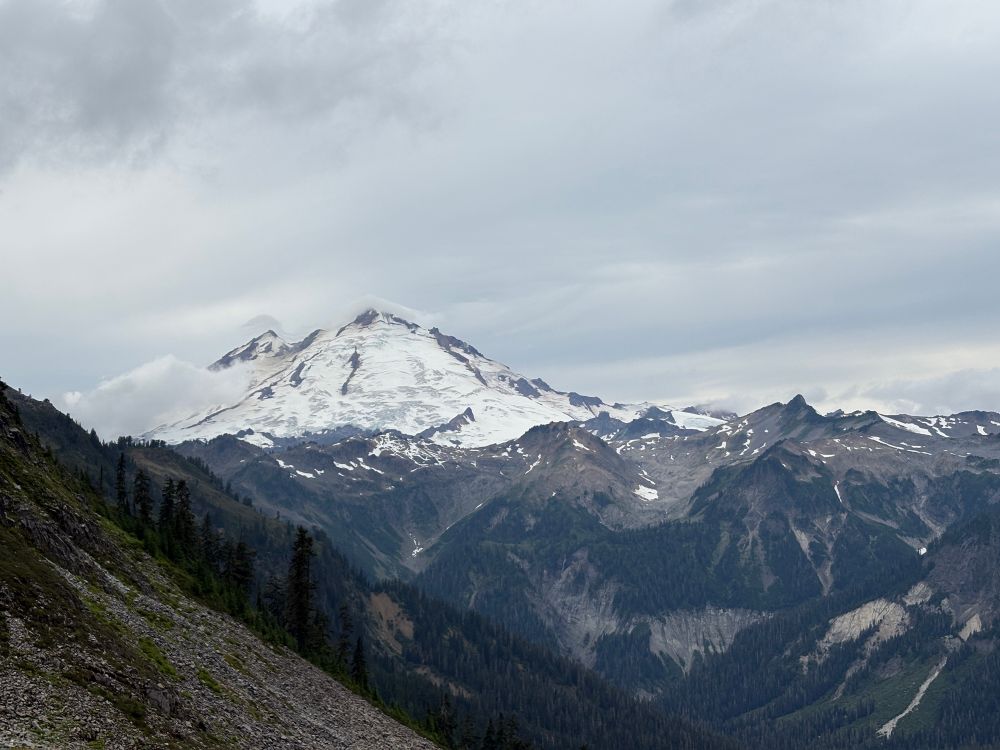 Photograph of a snow covered mountain peak with trees and rocky cliffs in the foreground 