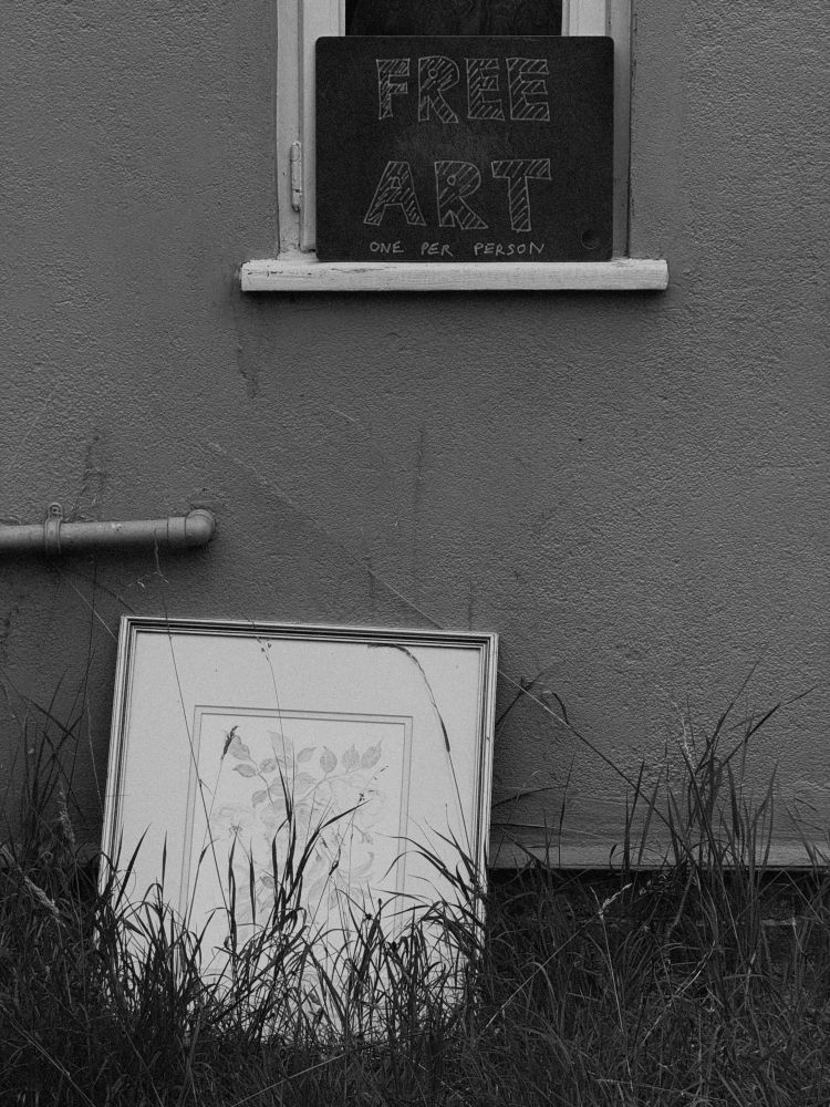 An old, framed picture sits among long grass and leant against a wall. On the windowsill above a sign has been written on a piece of slate. The sign reads “FREE ART, one per person.”