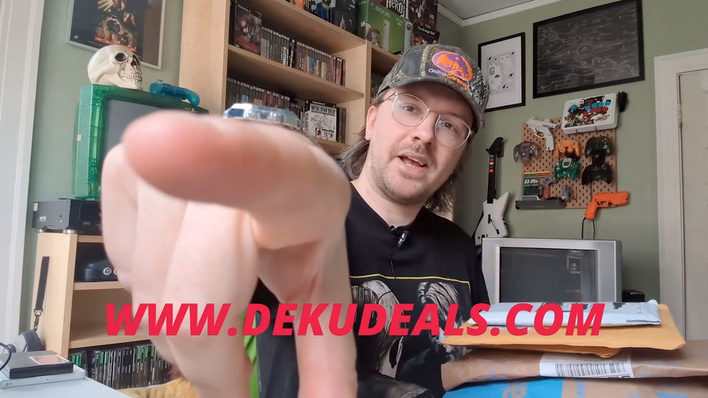 screen grab from Collector John's video on YouTube: https://youtu.be/vUhQ51dyjuI

A young person in glasses and a camo cap is holding a stack of packages and pointing at the camera. The letters "WWW.DEKUDEALS.COM" are shown across the image in bold red font.