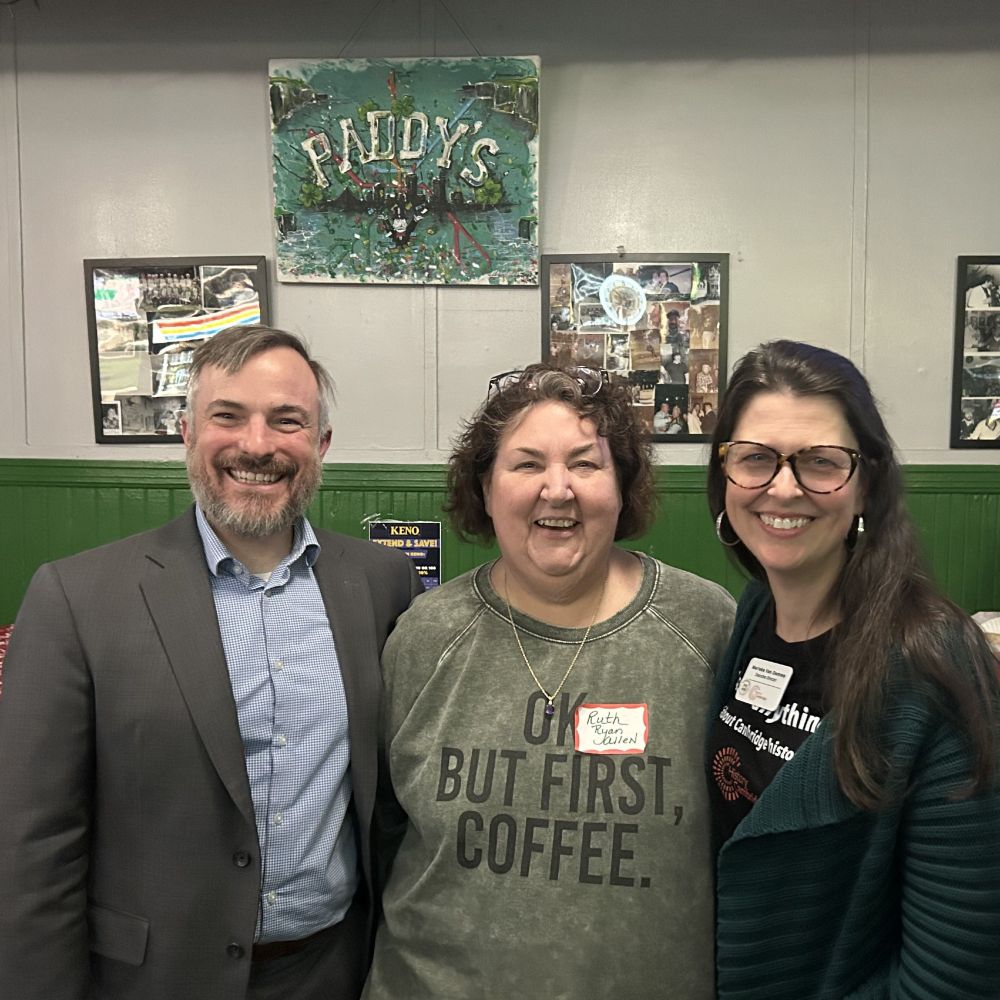 Three people standing next to each other smiling at the camera. The wall is half dark green (lower) and have off white (upper). On upper wall are framed photos and a handmade sign that reads Paddy's.