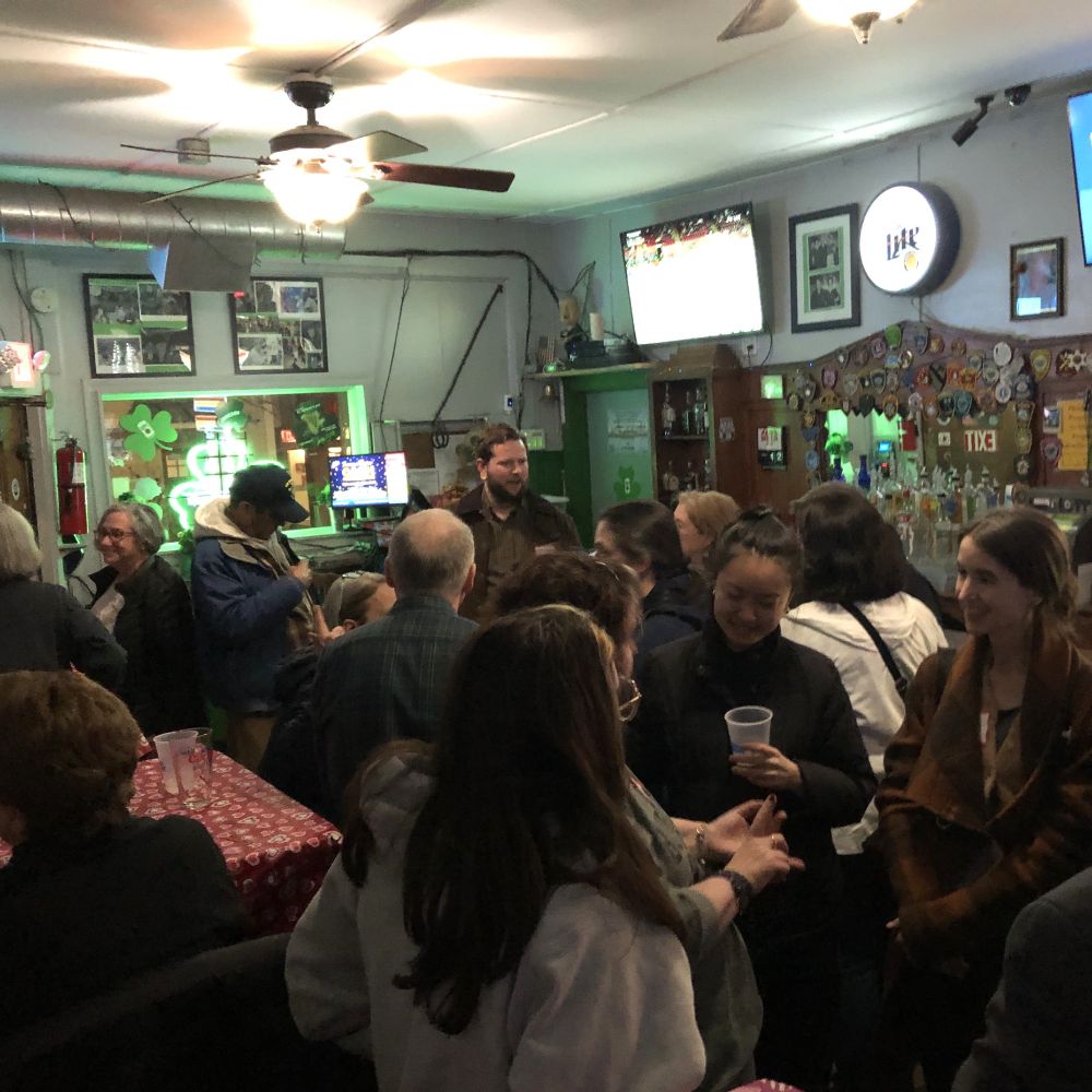 A dark crowded room of people talking. On the walls are framed photos, buttons, and cut out shamrocks. Person in the foreground is holding a cup.