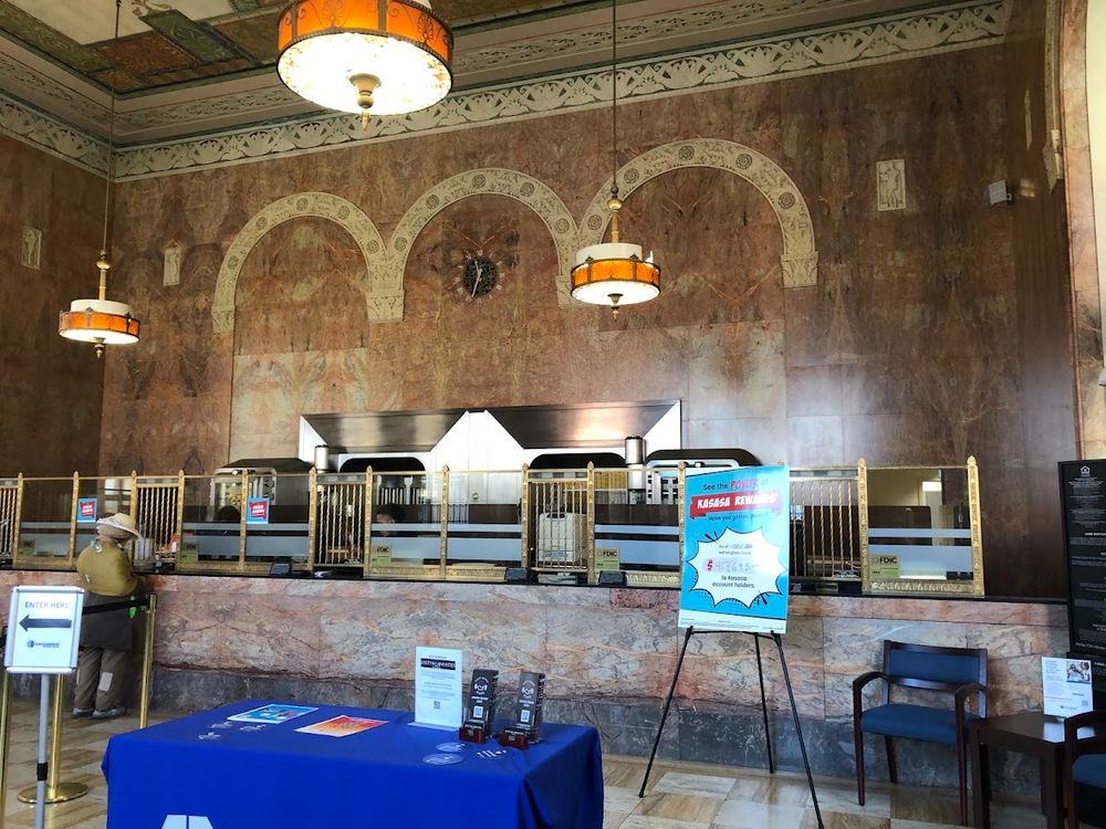 The grand interior of East Cambridge Savings Bank's main branch, showing the tall, marble-clad wall behind the teller line with decorative arched insets. Two large, ornate hanging lamps illuminate the space, and a table with a blue tablecloth and a sign are set up in the foreground.