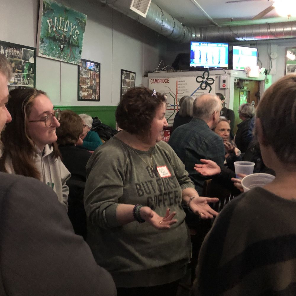 Woman with dark hair and green shirt is holding out her hands. She is in conversation with a person out of frame. The room is crowded with poeple talking to each other. A handmade sign on the wall reads Paddy's