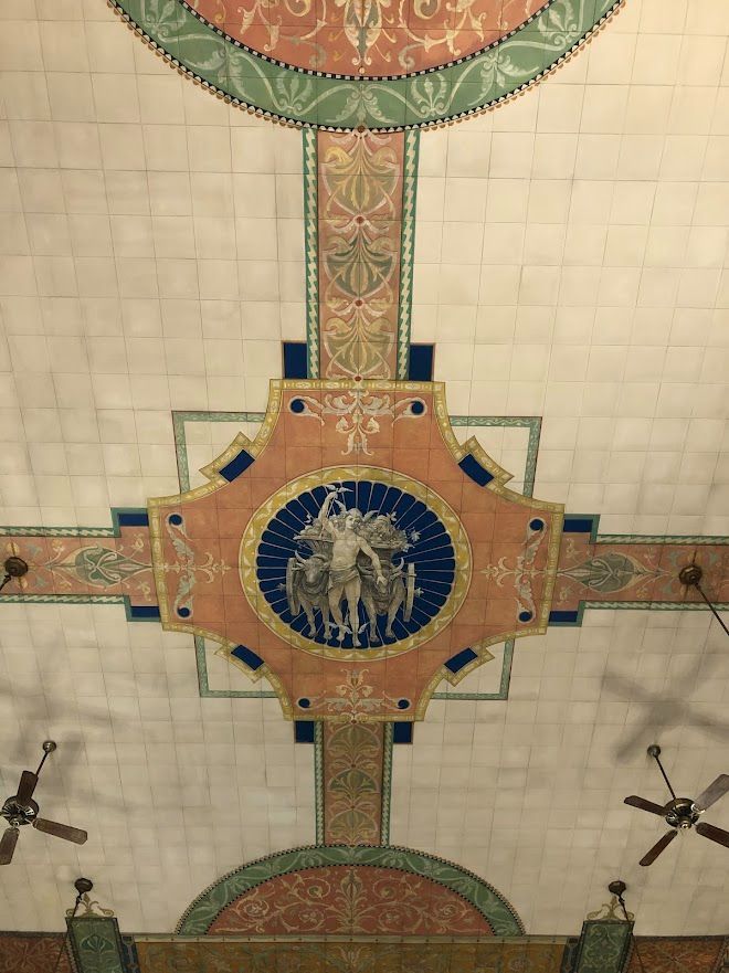 A detailed view of the ornate, painted ceiling inside the East Cambridge Savings Bank, featuring a central medallion with classical figures surrounded by intricate green, pink, and gold geometric and floral patterns. Two ceiling fans are visible in the corners.