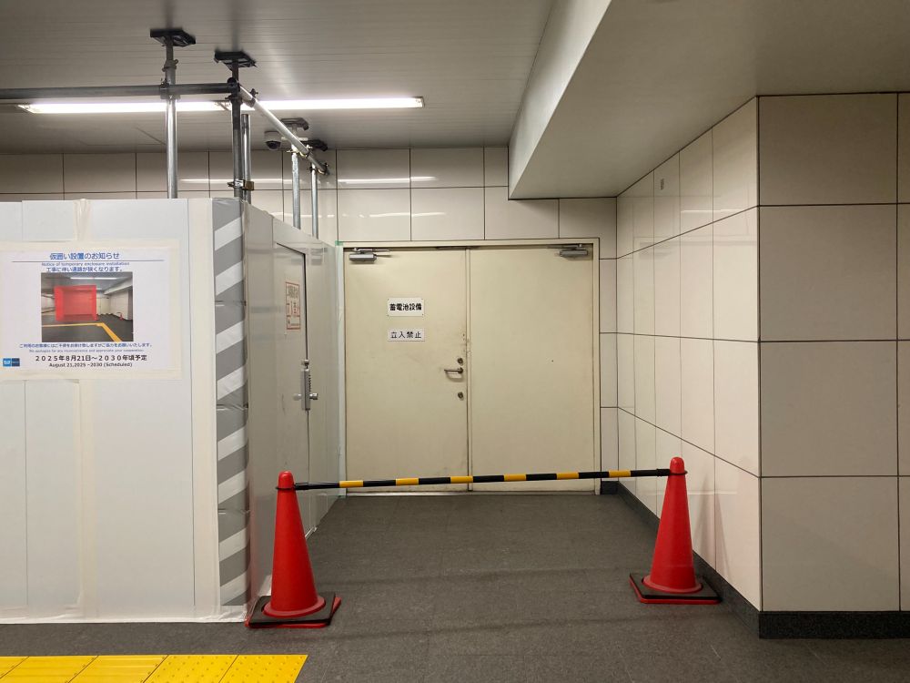 A corridor with a closed door and construction barriers. There is a notice about construction activities, and two orange cones are placed on either side of a yellow-and-black striped barrier. The walls have white tiles, and the area appears to be well-lit.