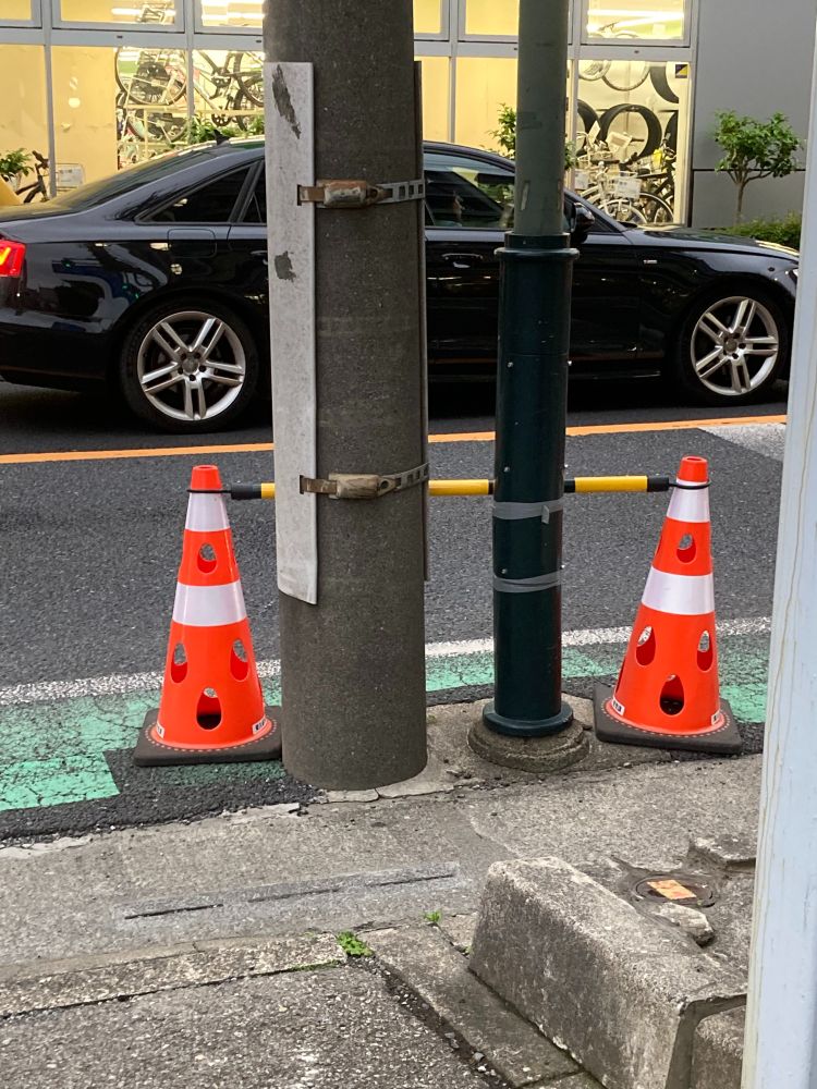 A black car is parked next to a traffic pole. On the street, there are three orange traffic cones positioned near the pole and a green bicycle lane. In the background, a window displays bicycles inside a store.
