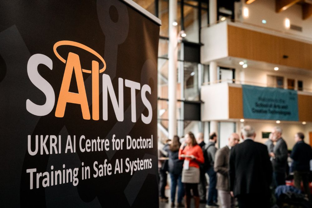 Banner with text: UKRI AI Centre for Doctoral Training in Safe Artificial Intelligence Systems (SAINTS).
There are a crowd of people in the background and out of focus. 