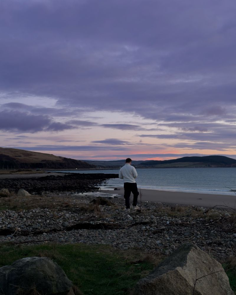 A person wearing a white sweater and dark pants stands on a rocky shoreline, looking out towards the ocean at sunset. The sky is filled with soft purple and pink hues, with scattered clouds. The coastline stretches into the distance, with rolling hills and a sandy beach meeting the calm waves. Large rocks and patches of grass are in the foreground, adding texture to the rugged landscape.