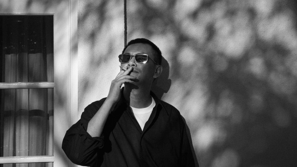 Black and white photo of Hong Kong film director Wong Kar-Wai wearing sunglasses and leaning against a wall while smoking a cigarette.