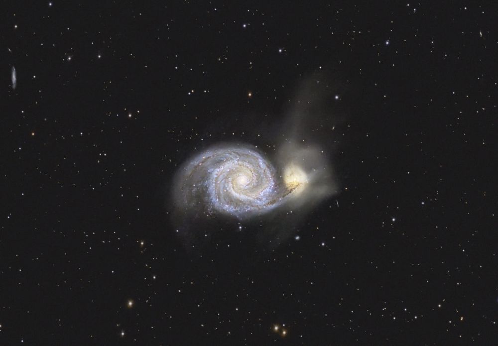 Two interacting galaxies glow in the colors of young and hot blueish stars, the yellowish tone of old and mature stars and exhibit magenta colored blobs of of excited hydrogen atom nebulae.