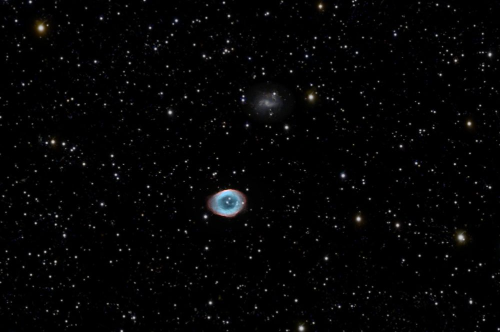 In front of a dark sky colorful stars shine together with the blueish-green-red gas remanants of a dying dwarf star. The scene is decorated with a tine spiral galaxy in the far distance. 