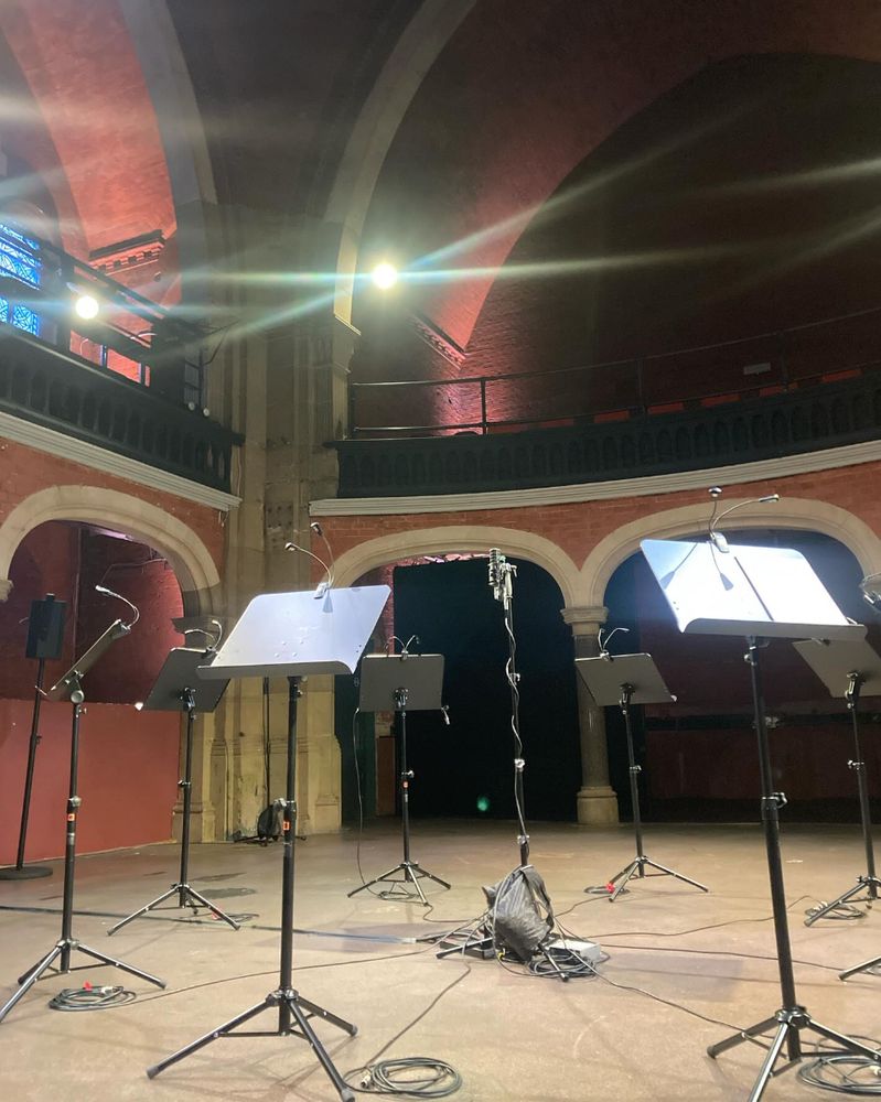 Music stands surrounding a central microphone in an old Victorian chapel - there are lights shining down on the music stands. 