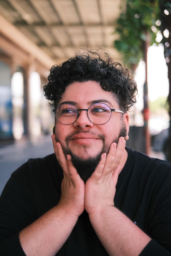 augie, a white trans masc person with short curly hair & round glasses, is looking off to the side. they've propped their chin up in both hands, which are covering most of their dark beard. they're smiling slightly