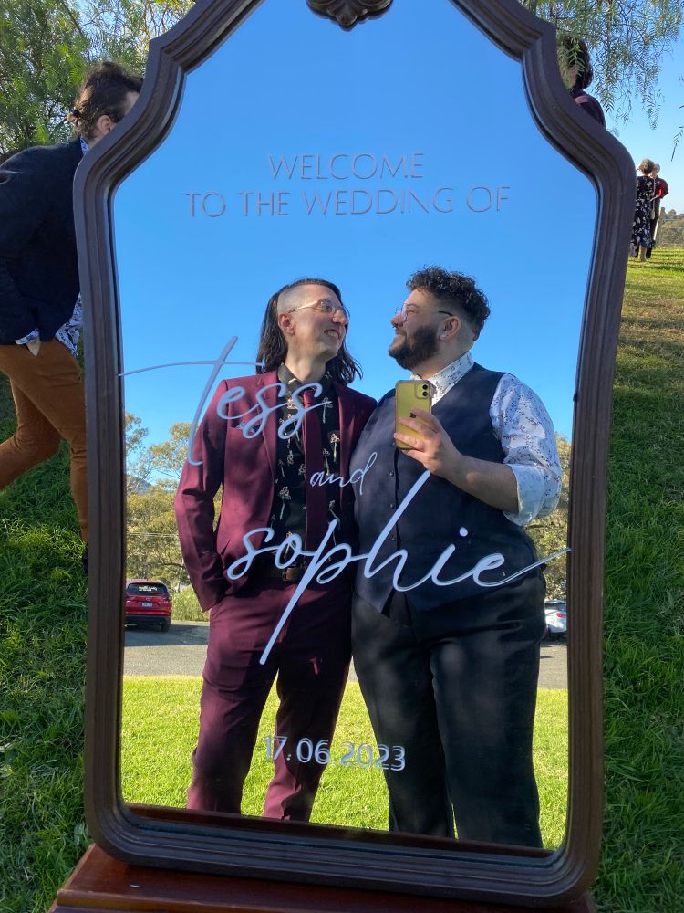 james and augie are reflected in a large mirror that says “welcome to the wedding of tess and sophie 17.06.2023”. james is wearing a burgundy suit and tie with his hair down and undercut facing the mirror, and augie is wearing a navy waistcoat and a white patterned shirt with rolled up sleeves. they’re looking at each other and smiling