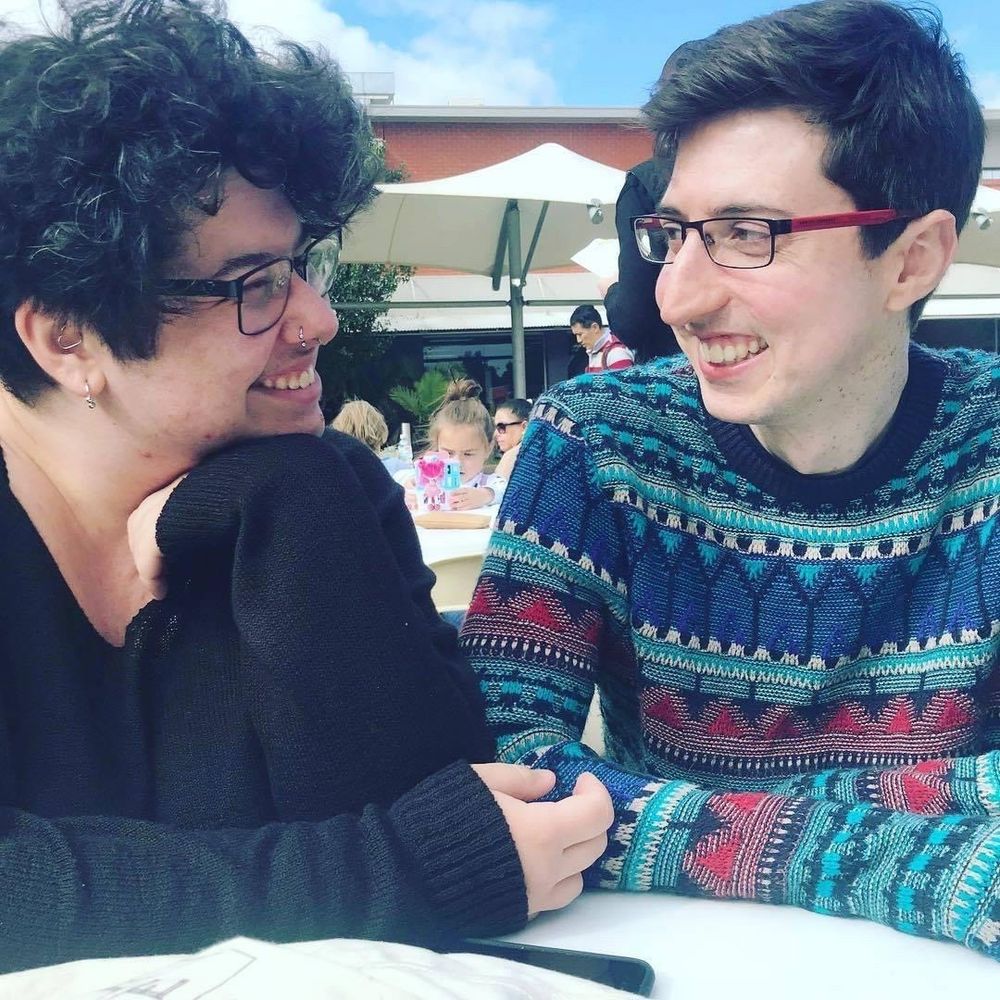 james and augie are sitting at a table at a market. they both have short hair and augie hasn’t grown a beard yet. they’re looking at each other and smiling