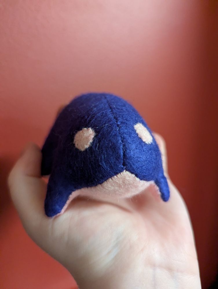 A photo of a small plush frog that fits in the palm of a hand. It is very simple and cute, with tiny legs and big round eyes