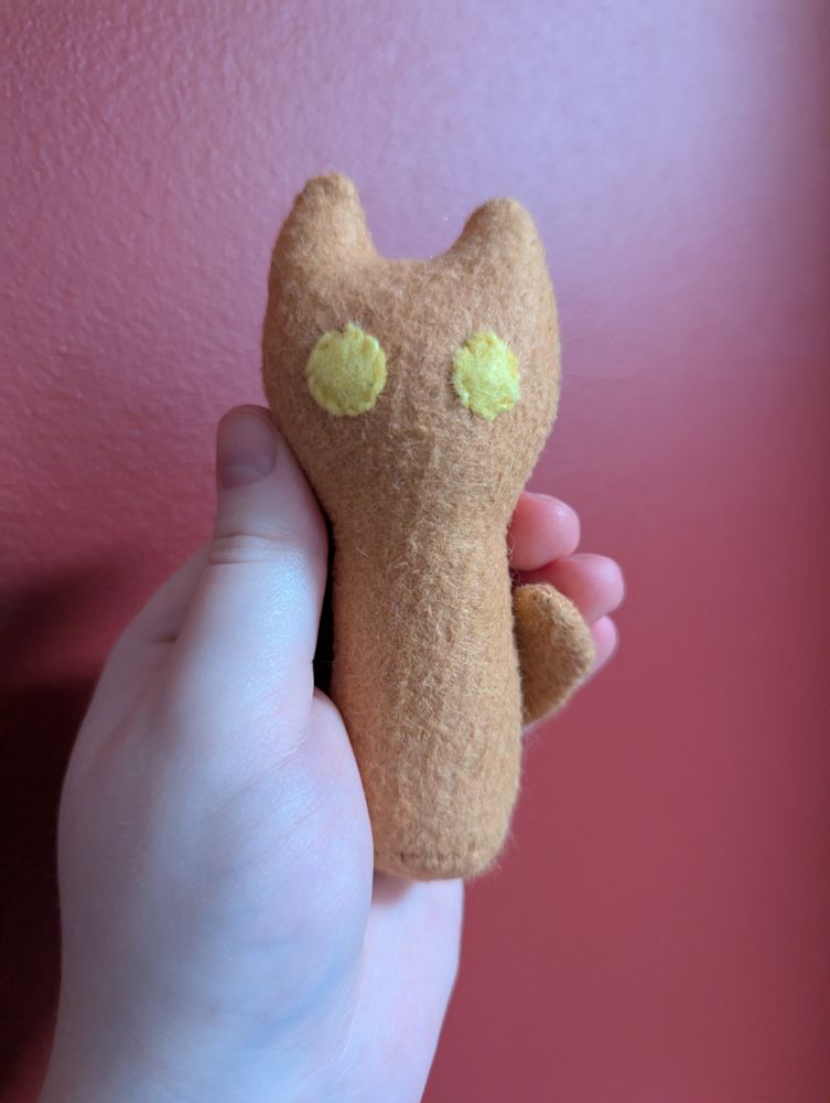 A photo of a small plush cat that fits in a hand. It is a simple shape of a cat, with pointed ears and no limbs besides a tail. It has big round eyes and no other features.