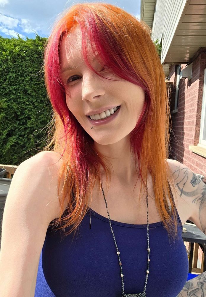 Ruby Soho takes a backyard selfie. She gives you a happy grin. Her hair is bright gingery orange with magenta streaks near her face. She wears a beaded necklace. A tattooed arm, just out of the picture, plays with her hair. Bright blue sky with some clouds, and a wooden fence are in the background behind her.