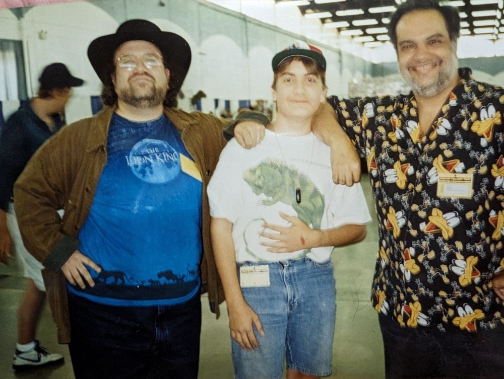two awesome comics creators flanking a scrawny nerd (me) dressed in terrible 90s denim shorts and a baggy tee. Peter David is on the left, sporting a black brimmed hat, hand on hip. George Perez is on the right, beaming a huge smile and wearing a handmade (by Mrs. Perez) button up shirt covered in Daffy Ducks. (Daffys Ducks?) 