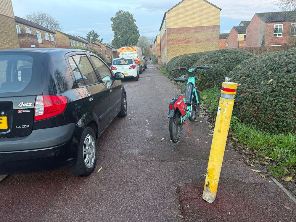 A yellow bollard placed in red tarmac called a Redway (cycle and foot path). There’s pavement parking on the Redway where cars and vans partially block the path. There’s also a parked hire bike on a stand blocking the way.