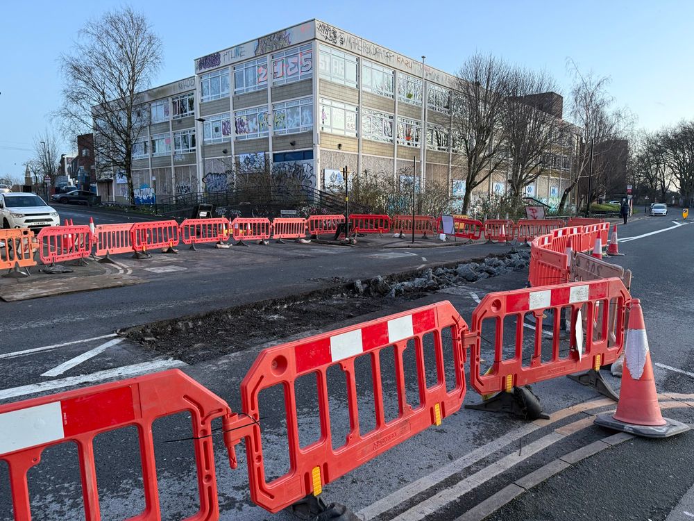 Roadworks at a crossroads. Part of the tarmac has been drilled into. There are many orange temporary barriers surrounding the hole in the road.