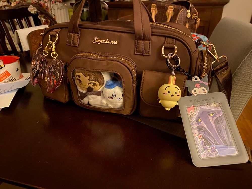 Brown bag on table, it is decorated with various keychains, and there is a pocket with a window showcasing two tiny plushies