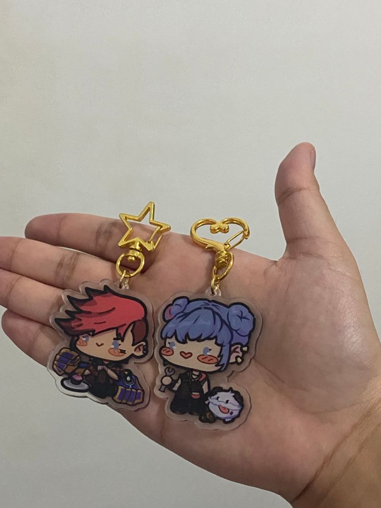 Keychains of Alternate Universe Powder and Canon Universe Vi from Arcane / League of Legends. The Vi keychain has a gold star-shaped chai, while the Powder keychain has a gold heart-shaped chain.