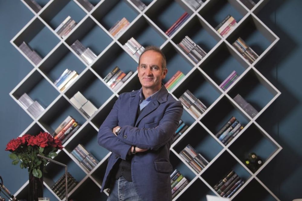 Kevin McCloud from Grand Designs stands in front of a geometric bookshelf 