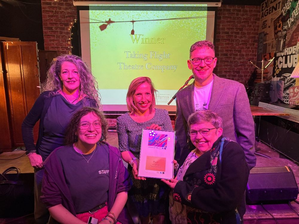 Louise, Steph, Elise, Alan and Sara pose with the award, which is a framed certificate. On a screen behind them the words Winner Taking Flight are visible on a yellow background. The lighting is very hot pink in tone and everyone is smiling. 