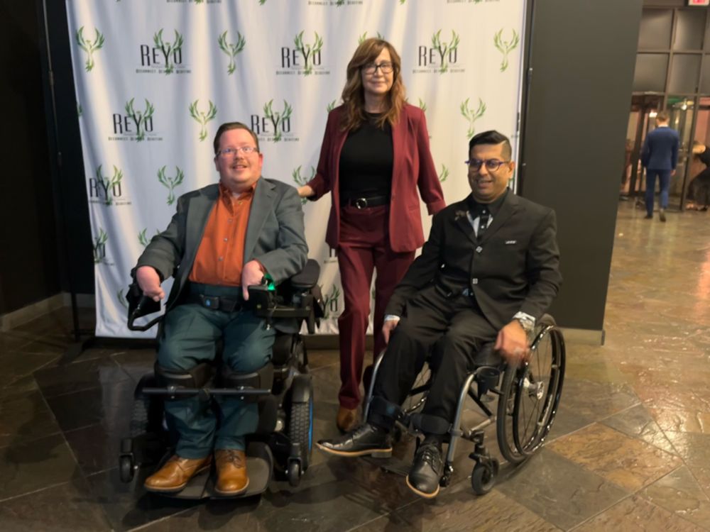Zachary, Marie, Aleem together with the Reyu backdrop. Two are in their chairs. 