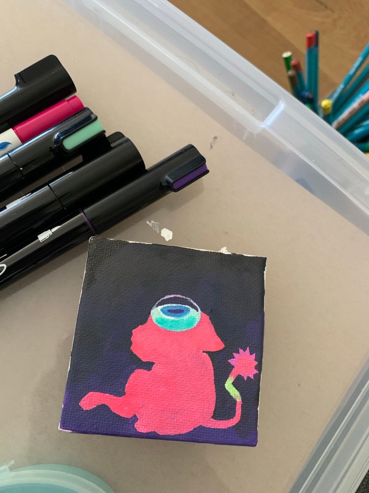 Work in progress painting on a small canvas showing a pink creature with a giant eye looking up. The canvas is surrounded by a few acrylic paint markers and is resting on a clear art material box.