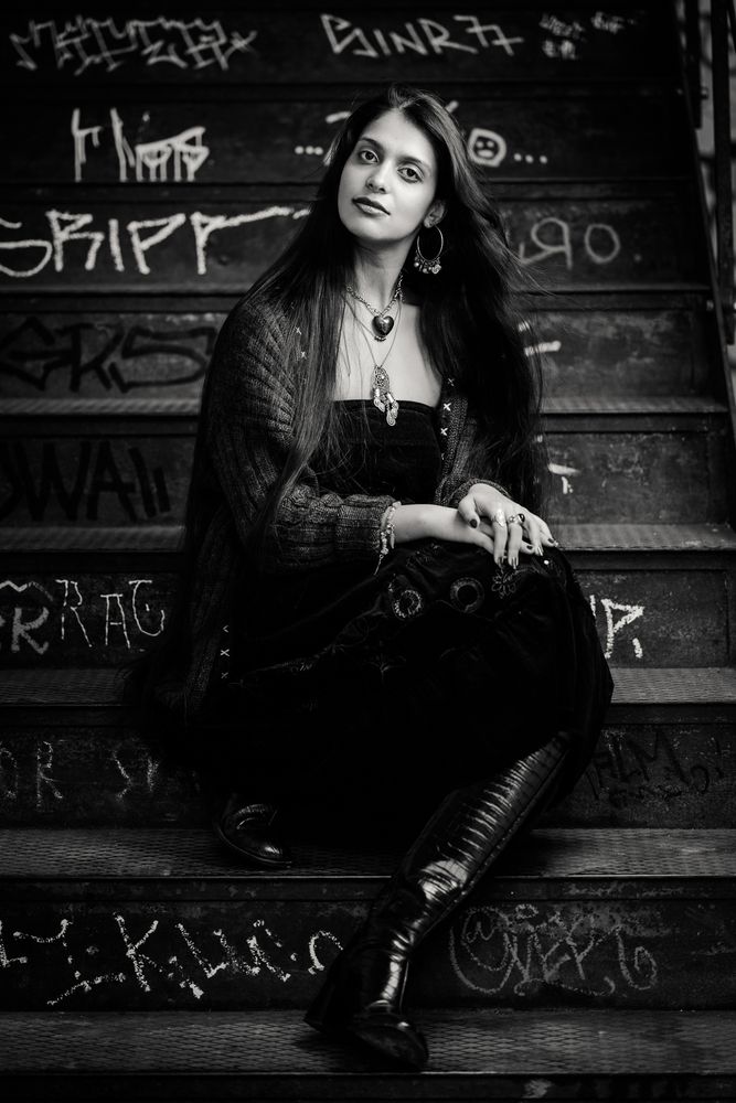 The black and white portrait shows a woman seated on a weathered, graffiti-covered metal staircase. She wears a black strapless top under a dark cardigan, a skirt with embroidery, knee-high leather boots, rings, a bracelet, hoop earrings, and layered necklaces. Her long dark hair frames a pensive expression as she gazes upward. The soft lighting and urban backdrop create a moody, artistic atmosphere with a vintage feel.