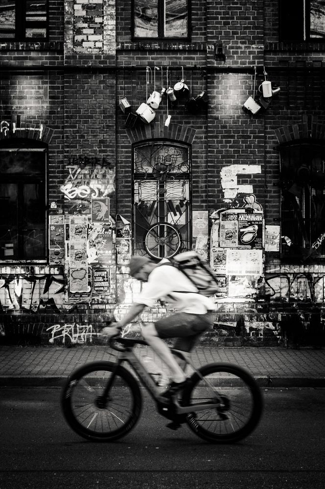 A cyclist is the primary focus of this black and white image. He is seen in motion, riding along a street with a weathered brick building as the backdrop. Graffiti and tattered posters adorn the walls, hinting at an urban, artistic atmosphere. Hanging objects give a sense of the building's history and the city's alternative scene.