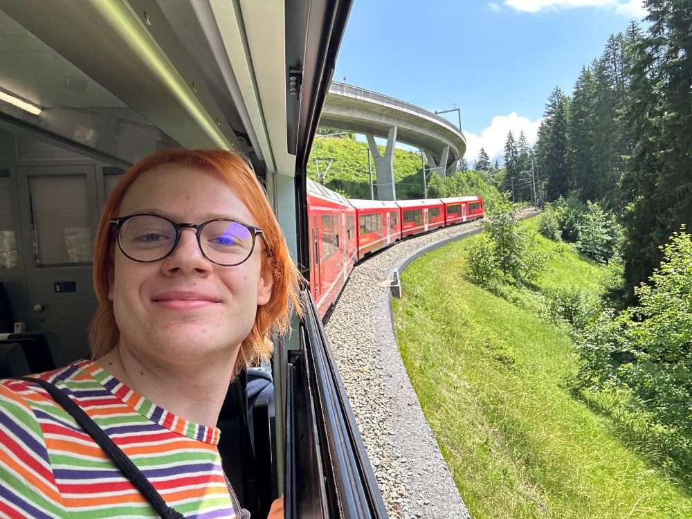 I am standing next to an open window of an RhB Capricorn. The scenery I’m surrounded by is rather alpine.