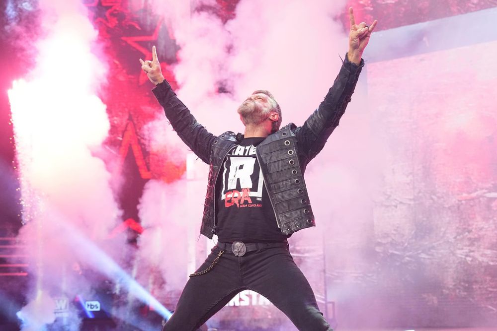 Image of AEW wrestler Adam Copeland during his entrance to Altar Bridge’s Metalingus. He is standing with his legs spread and his arms raised, with his fingers in the rock “horns” sign 🤘. He is wearing black jeans, a black jacket & a tshirt stating “Rated R Era”. Smoke billows behind him.