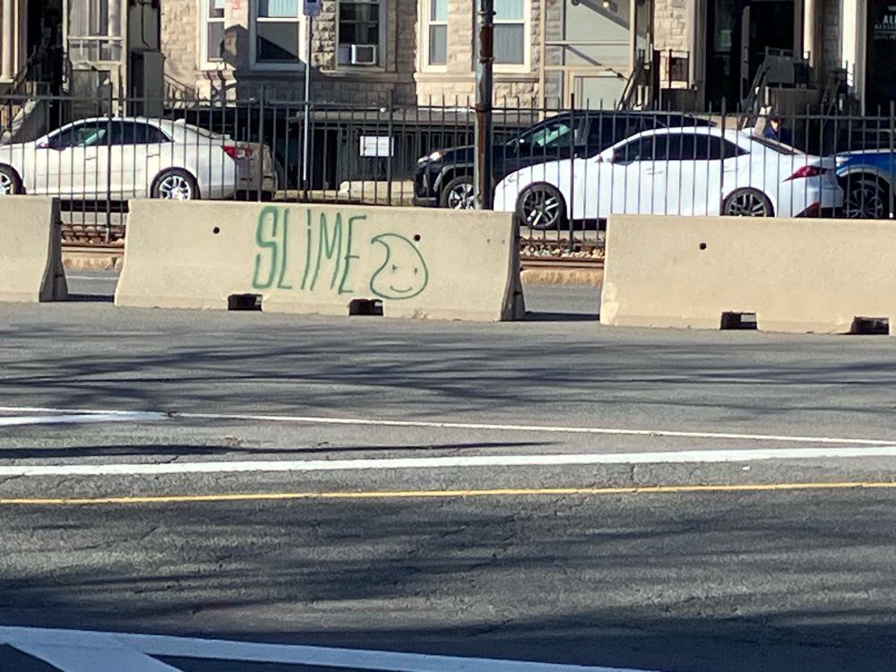 The word “SLIME” and a little picture of a dragon quest slime spray painted in green on a street divider