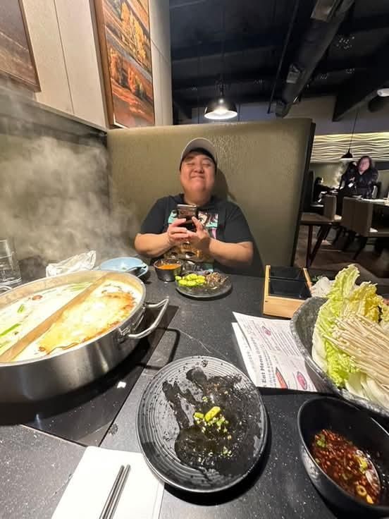 Nico, a fat brown queer is sitting and smiling eating Hot Pot at Happy Lamb 