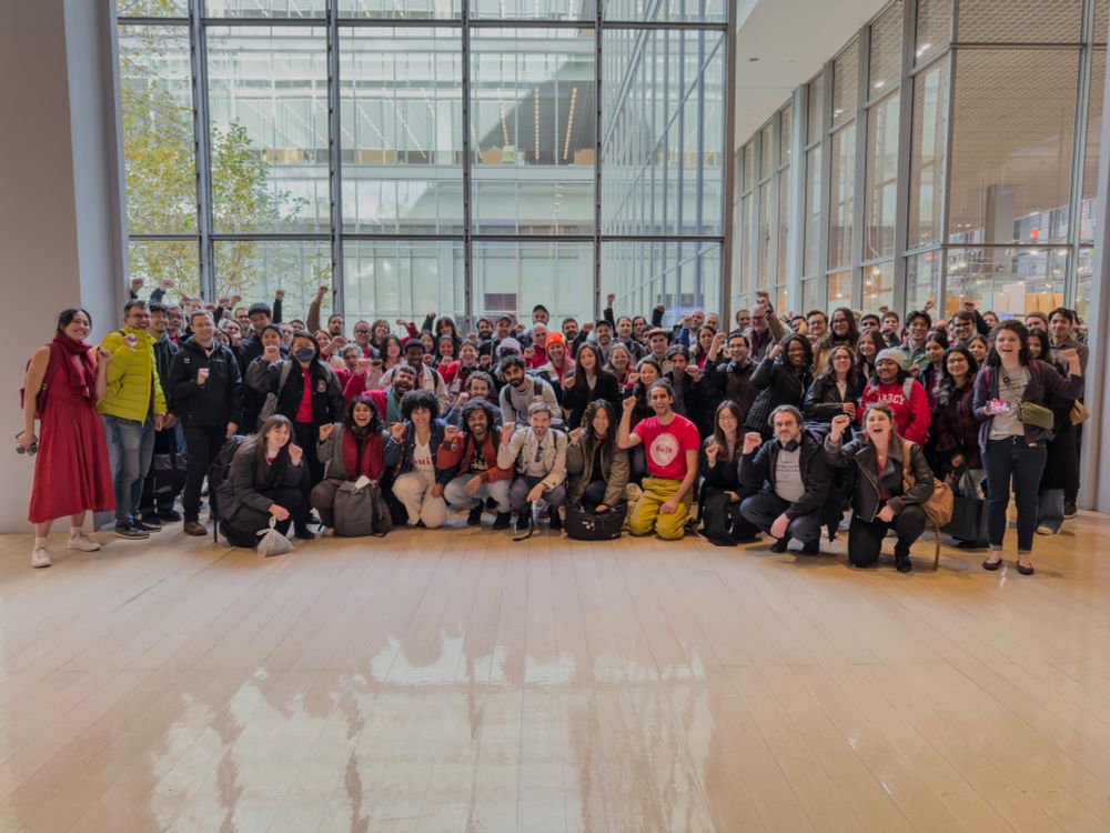 A group photo of about a hundred members of the Times Tech Guild, standing in the lobby of the New York Times building, many with fists raised in solidarity. In front of them is a shiny light wood floor, and behind are large floor to ceiling glass windows overlooking an inner greenery. Several members are wearing read dresses and shirts.