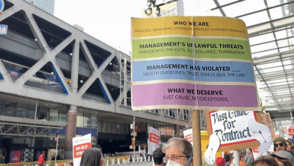 An animated GIF of the picket line in which a man wearing glasses and a mask carries a large sign with four colored rectangles on it past the camera. Other protest signs can be seen in the background. The large sign says: Who We Are - analysts, designers, engineers, product & project managers, in a yellow box. Management’s Unlawful Threats - forced RTO, pay cuts, surveillance, termination, in a green box. Management Has Violated - health guidelines, trust, status quo, the law, in a blue box. What We Deserve - just cause, no exceptions, in a purple box.