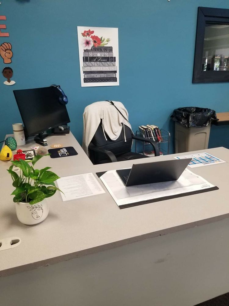 School teacher desk with laptop, calendar, and sensory toys that include a green dragon egg, zen garden, and stacking stones. There is a small philodendron plant. Neat and organized. 