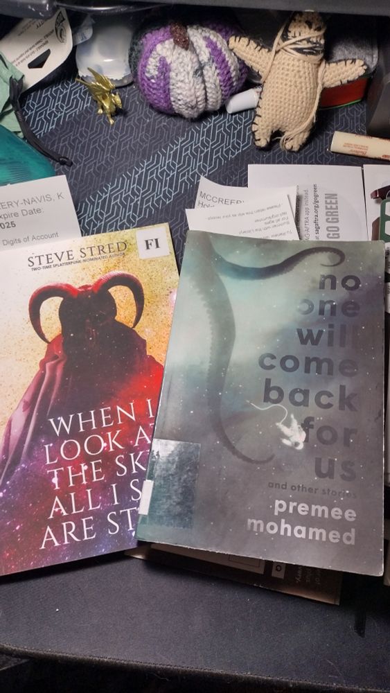 Photo of a cluttered desk with a crocheted voodoo doll and small  crocheted pumpkin in the colors of the non-binary flag and featuring the books "no one will come back for us" by Premee Mohamed and "when I look at the sky all I see are stars" by Steve Stred.