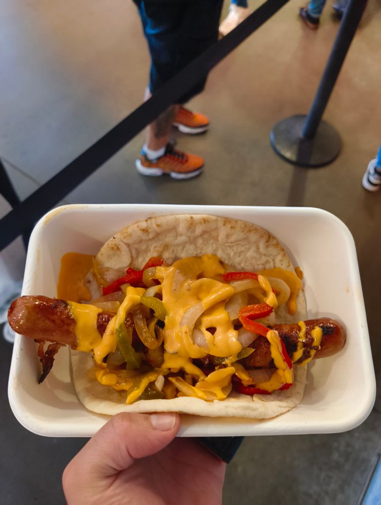 On a white tray sits a bacon wrapped hot dog, topped with assorted grilled onions, peppers & a yellow sauce, itself sitting atop a pita bread.