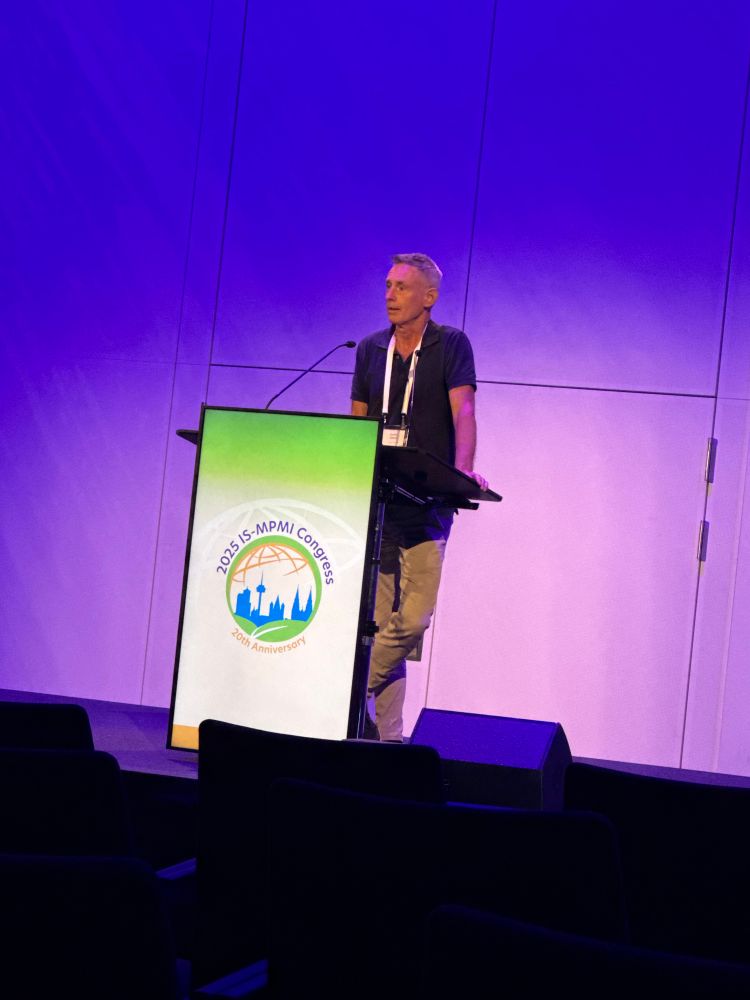 A man giving a talk at a lectern with the MPMI logo